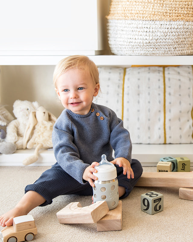 Toddler sitting and holding an 8 ounce stainless steel baby bottle with silicone nipple in hand.