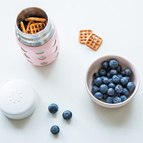 Lifefactory stainless steel baby bottle with pretzels inside, with flat cap next to it to show multiple uses.