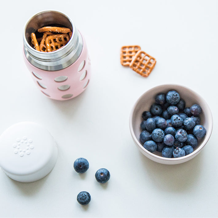Lifefactory stainless steel baby bottle with pretzels inside, with flat cap next to it to show multiple uses.