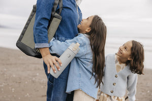 Two children with their mom on a beach.  Mom is holding a Lifefactory water bottle and has a grey wine tote over her shoulder. 