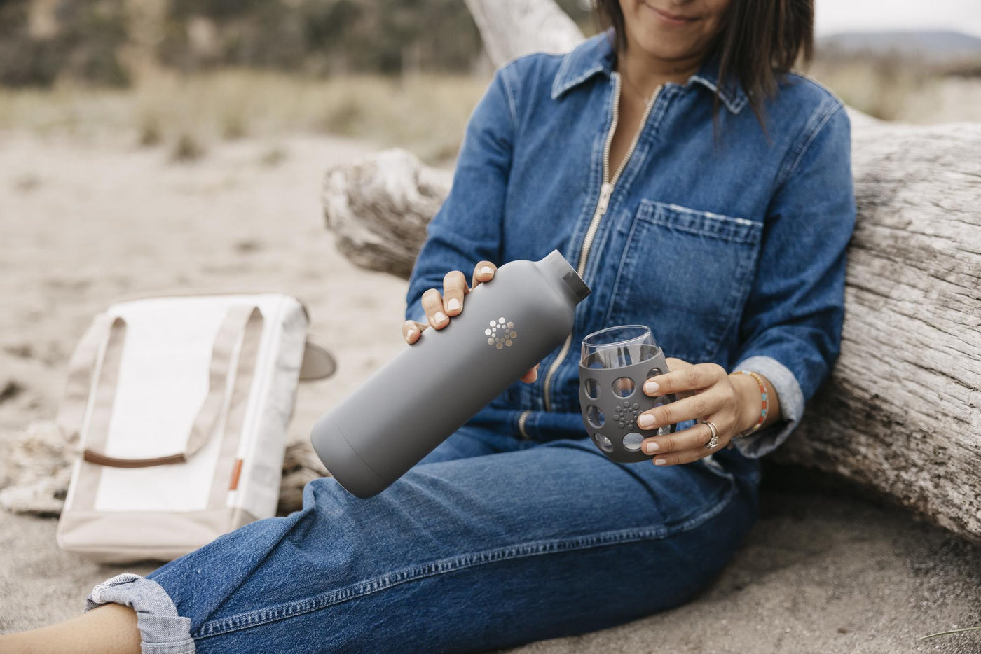 Woman sitting on a beach with a Lifefactory wine tote, using a Stainless Steel water bottle to pour a drink into a wine glass.