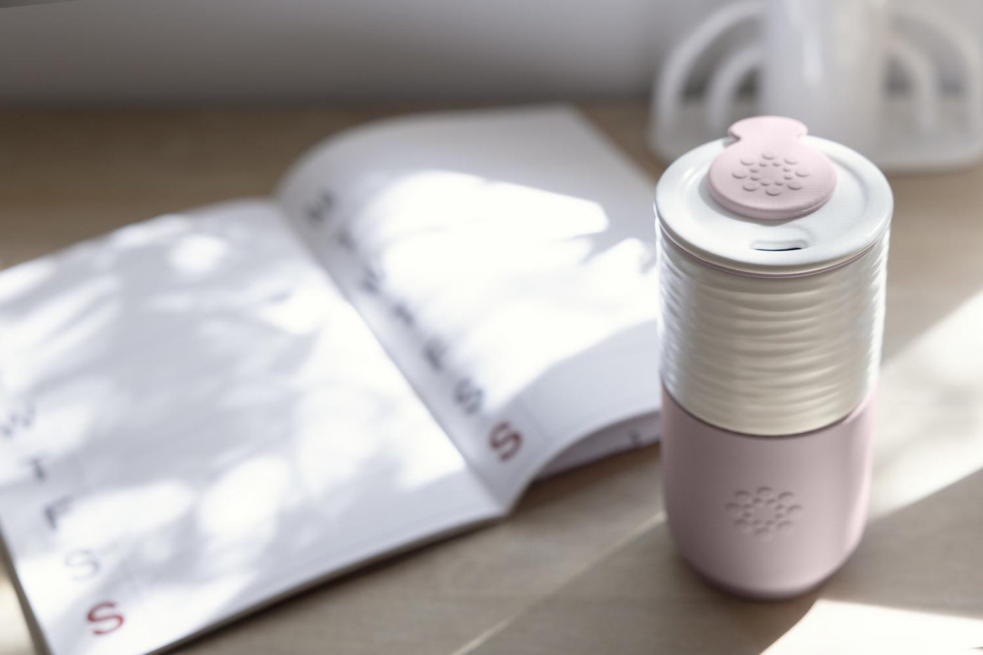 Lifefactory Pink and white ceramic travel tumbler with lid and silicone drink stopper on a wooden surface with an open book in the background