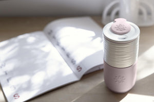 Lifefactory Pink and white ceramic travel tumbler with lid and silicone drink stopper on a wooden surface with an open book in the background