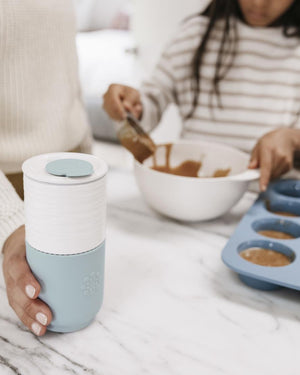 Person holding a Lifefactory white and blue ceramic tumbler with another person baking in the background.