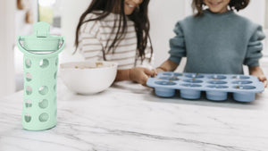 Two children in a kitchen setting with a Mint Lifefactory glass water bottle water bottle and cupcake tin on a marble countertop.